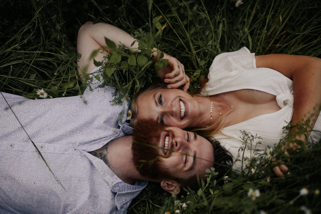 A couple is lying in the grass side by side for their engagement photos. They are looking up and smiling.
