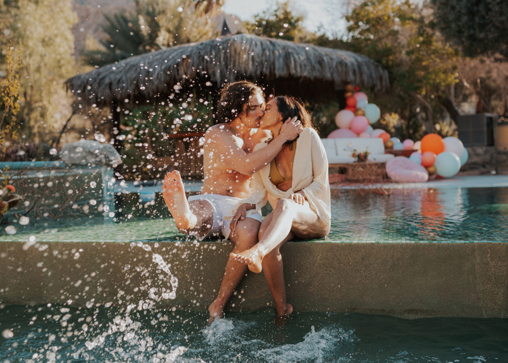 A couple is taking engagement photos by the pool. They are sitting side by side, kissing, with water splashing in front of them.