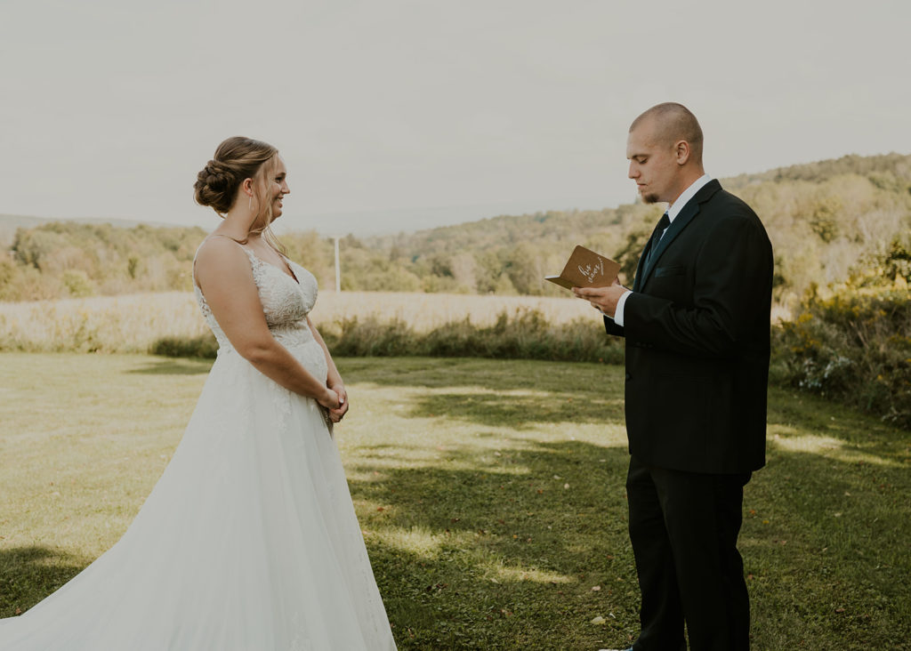 A couple is facing each other in front of a field of grass after their first look. The groom is reading from a booklet titled "his vows."