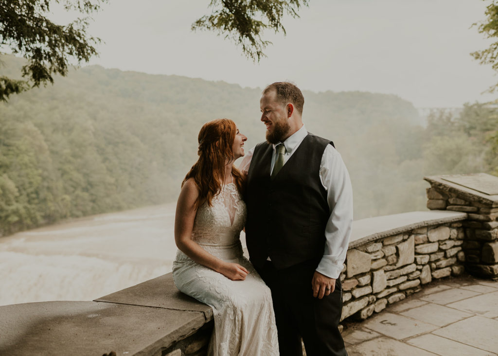 A bride is sitting on a stone wall in front of a waterfall at the Glen Iris Inn wedding venue in Livingston County. The groom is standing next to her, and they are looking at each other with their arms arund each other.