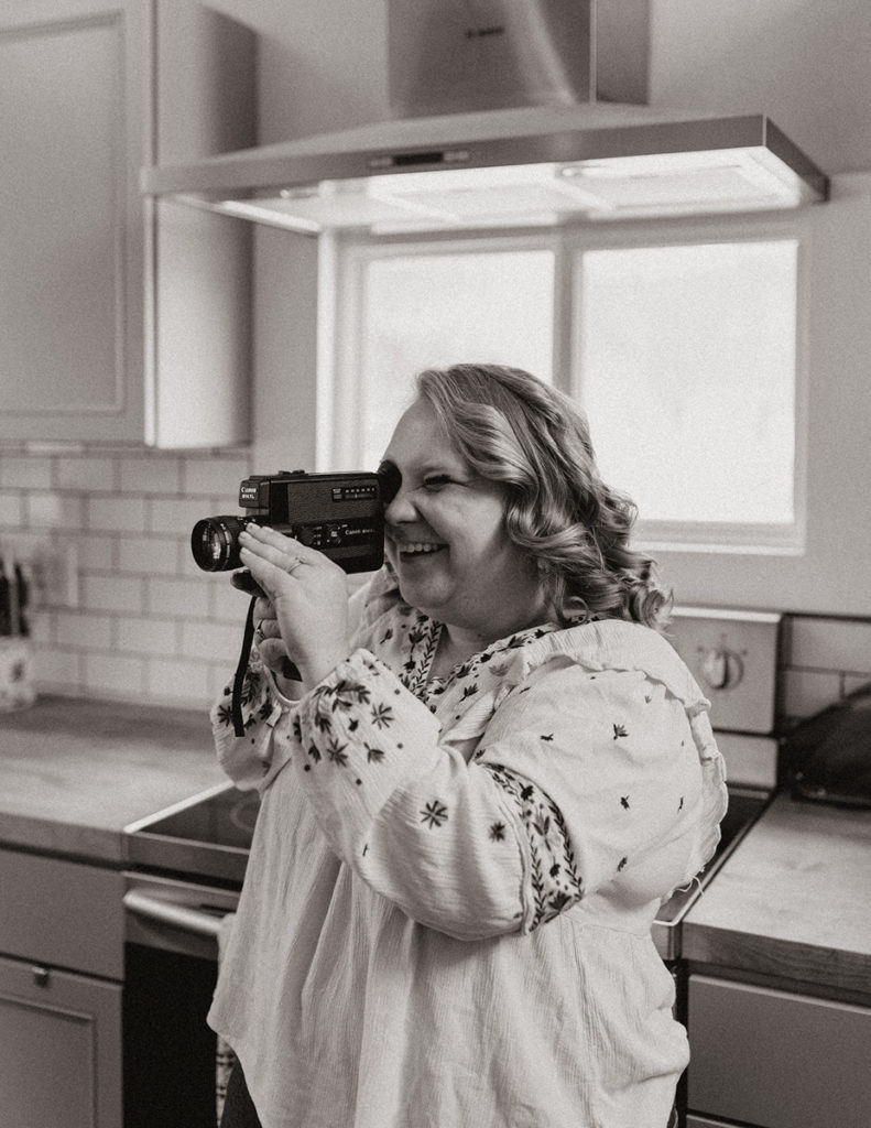 A black and white photo of a woman standing in a kitchen, holding a Super 8 film camera and smiling.