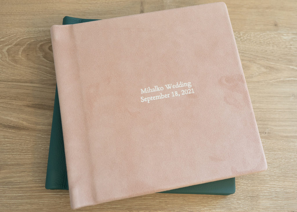 A pink wedding photo album is stacked on top of a green one, lying on a wooden desk.