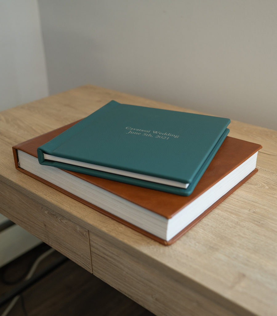 A green wedding photo album is stacked on top of a brown one, lying on a wooden desk.