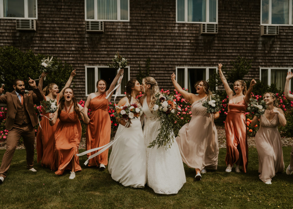 Two brides are kissing, surrounded by their wedding party after their first look.