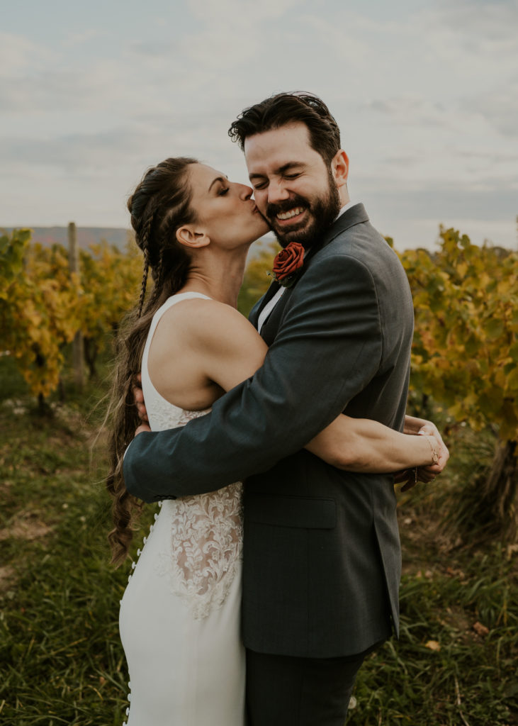 A couple hugging in a vineyard wedding venue, with the bride kissing her groom on the cheek.